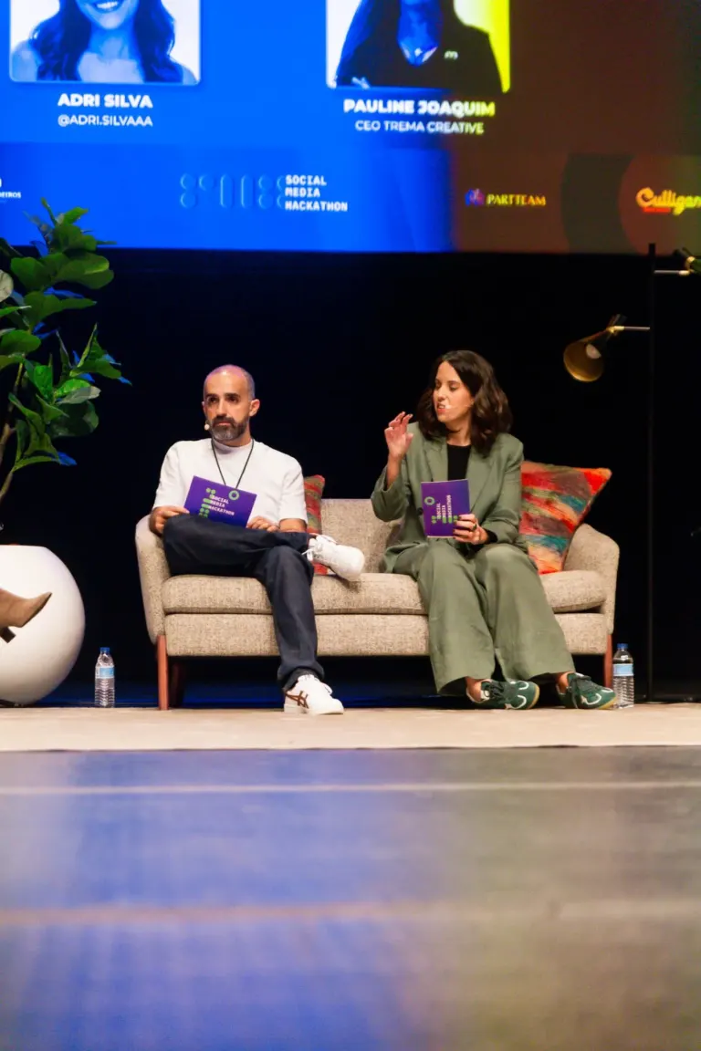 Dois palestrantes sentados em um sofá em um evento, com um pôster eletrônico ao fundo exibindo nomes e informações de...