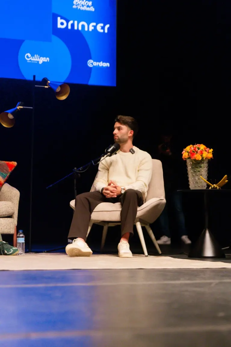 Homem sentado em cadeira branca, com cabelo castanho, barba, usando suéter branco, calça marrom e tênis brancos, em palco...