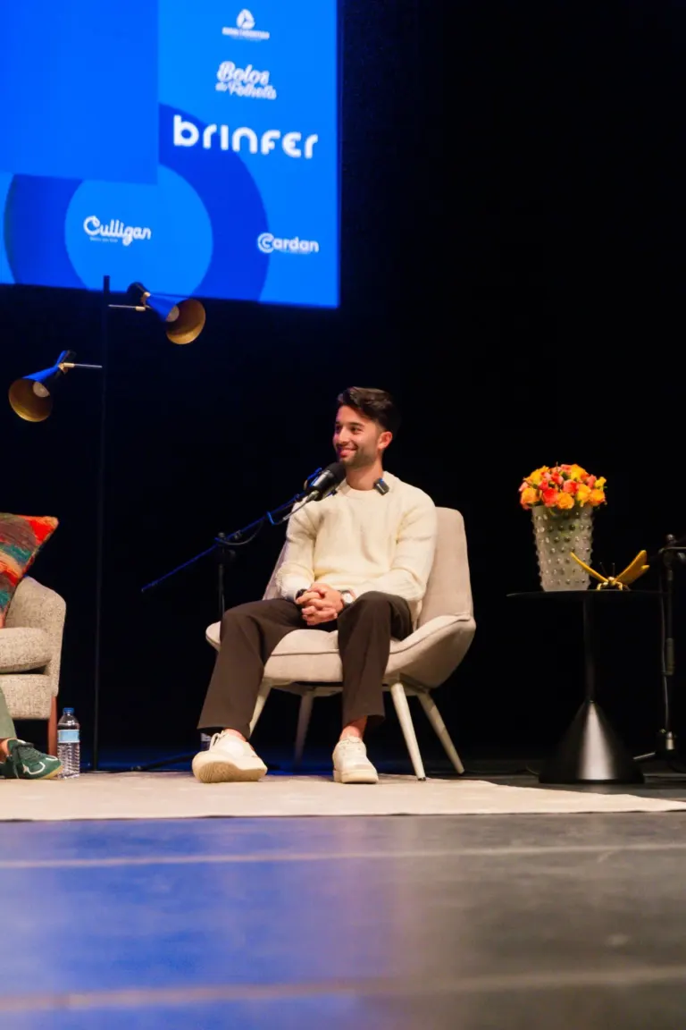 Homem sorridente sentado em uma cadeira em um palco com microfone, ao lado de uma mesa com vaso de flores, em evento com...