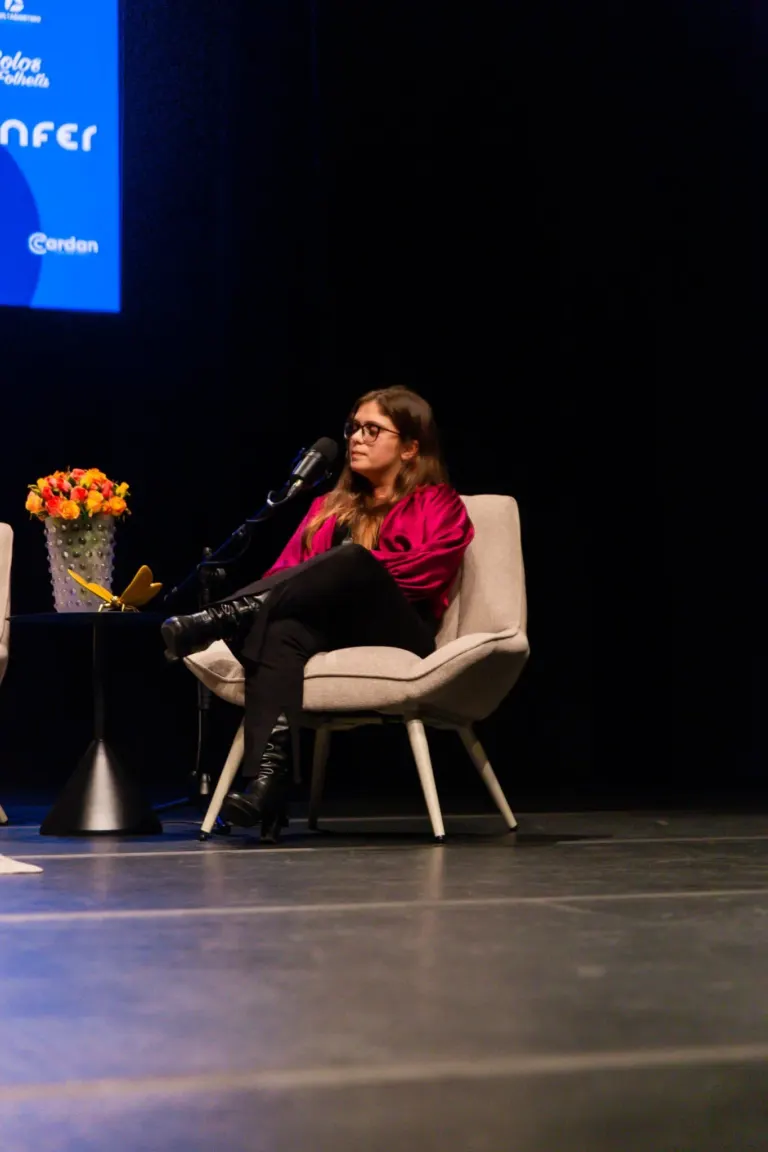 Pessoa de cabelos longos, usando óculos e roupa rosa, sentado em uma poltrona em um palco, com microfone diante dela, ao...
