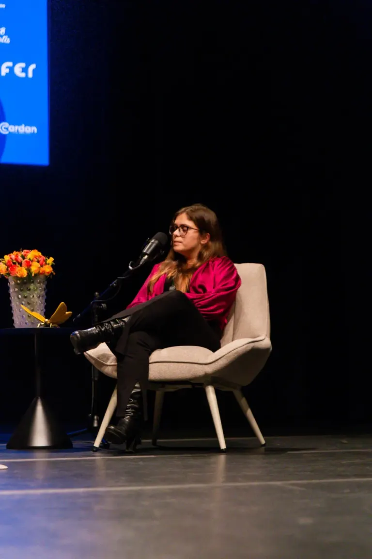Mulher de cabelo castanho, óculos, vestida com blusa rosa sentada em cadeira confortável em palco, falando ao microfone,...