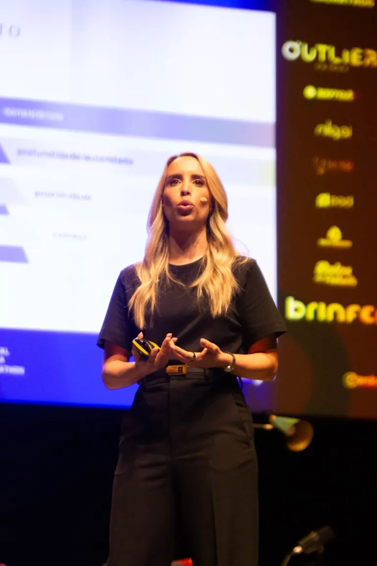 Mulher de cabelos loiros longo, vestindo roupa preta, falando em palco durante uma apresentação, com tela de fundo...