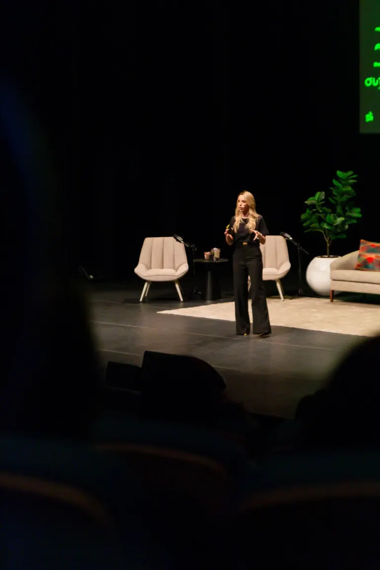Mulher de cabelo loiro apresentando em palco com móveis de sala decorativa, incluindo duas cadeiras, uma mesa com objetos...