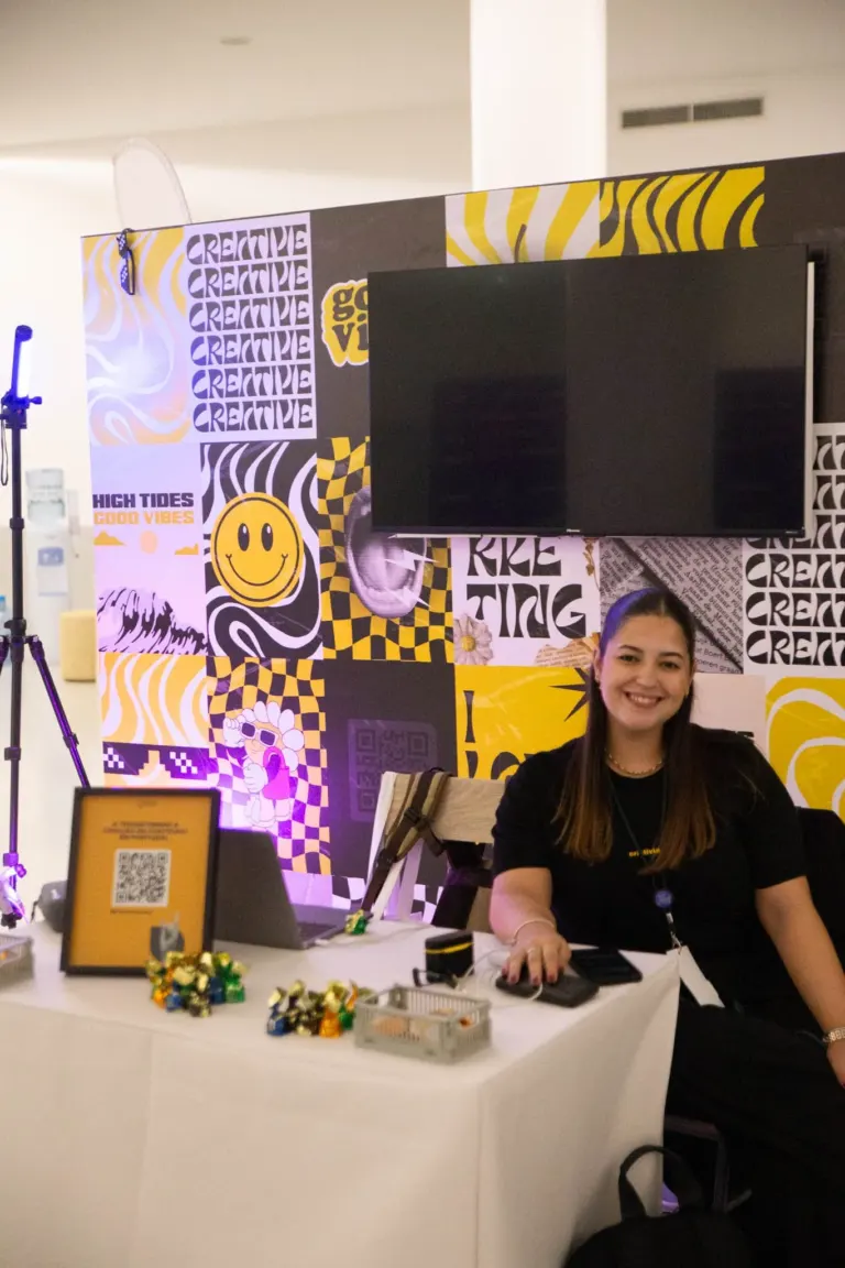 Pessoa sorrindo sentada atrás de uma mesa decorada com rapaduras de açúcar, com um fundo colorido de painéis com desenhos...
