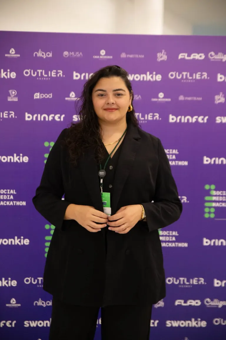Mulher com cabelo cacheado escuro, vestindo blazer preto e acessórios dourados, posando em frente a um fundo roxo com...