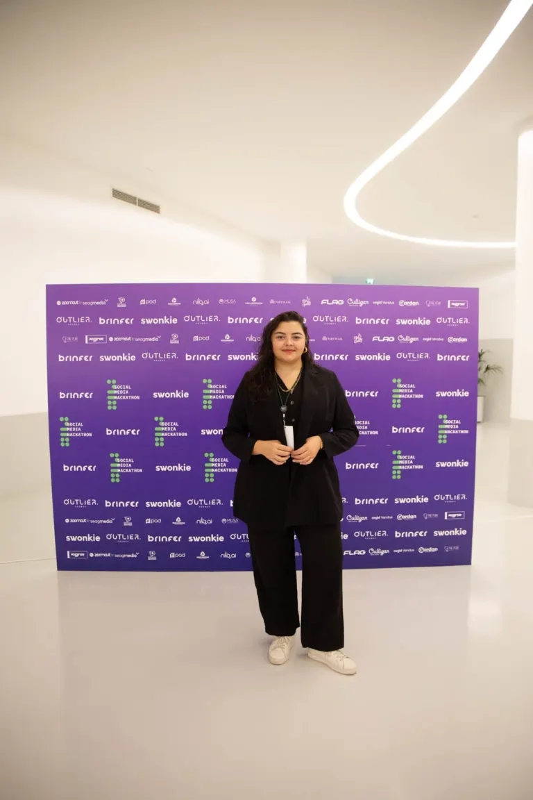 Mulher de cabelo escuro com roupa social preta e tênis branco posando diante de um painel roxo com logotipos de eventos e...