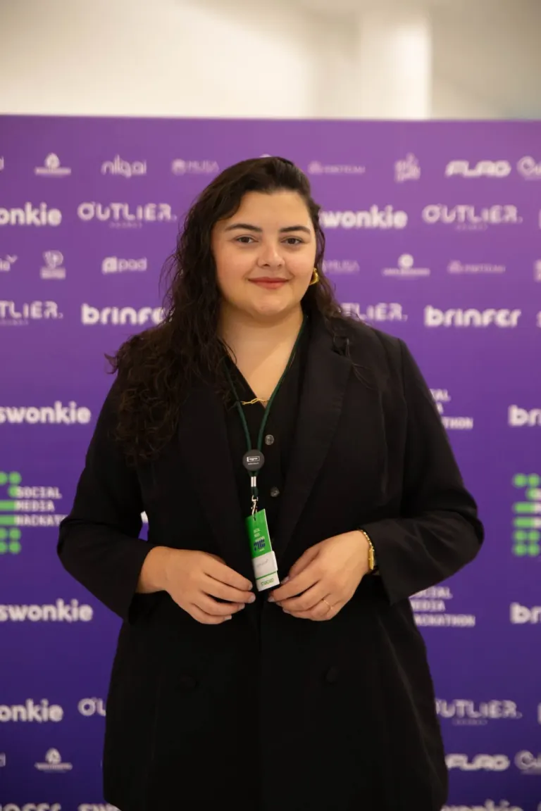 Mulher de cabelos castanhos escuros e pele clara, usando blazer preto, com crachá, em evento com fundo roxo com logotipos...