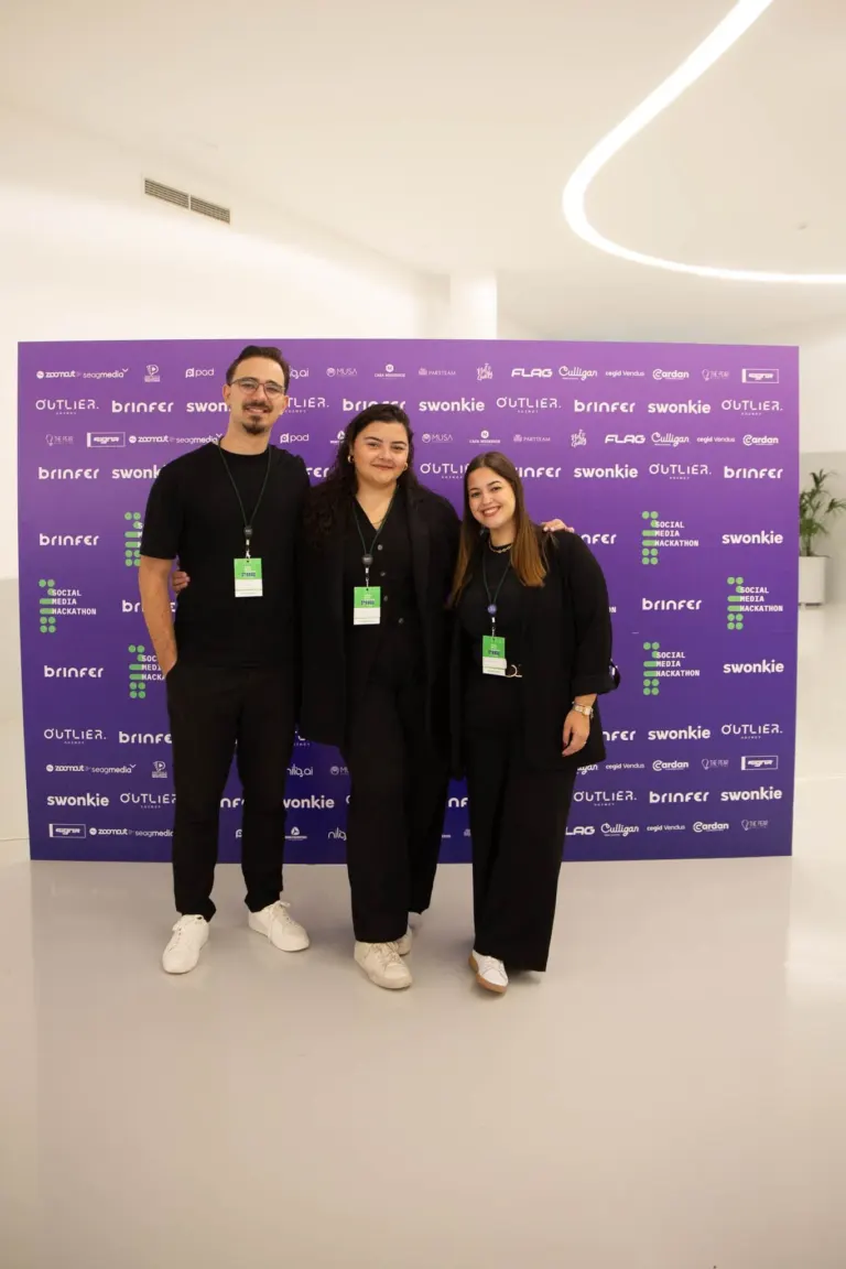Três pessoas sorrindo posando na frente de um painel roxo com logos de eventos e empresas, em um ambiente interno bem...