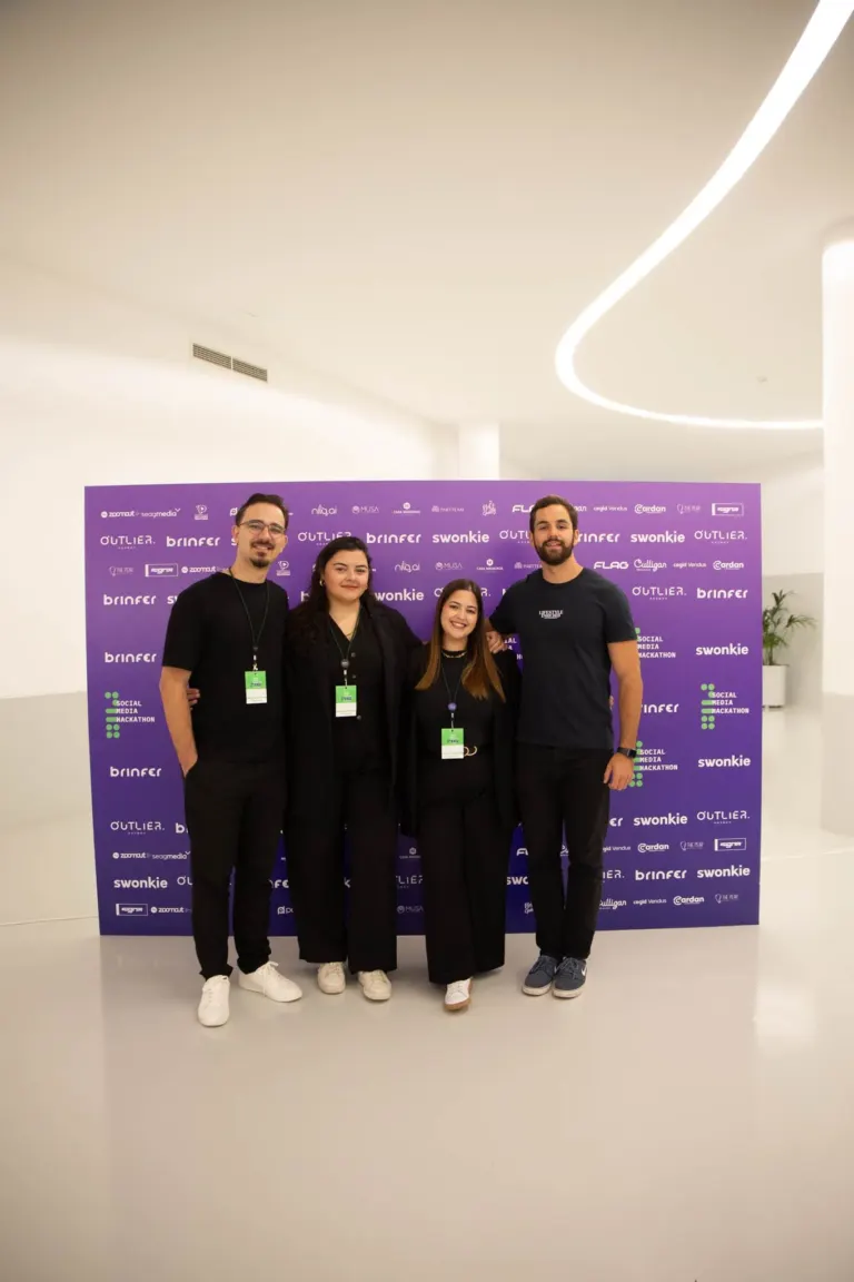 Grupo de quatro pessoas posando para a foto em frente a um painel roxo com logotipos e nomes de eventos e patrocinadores,...