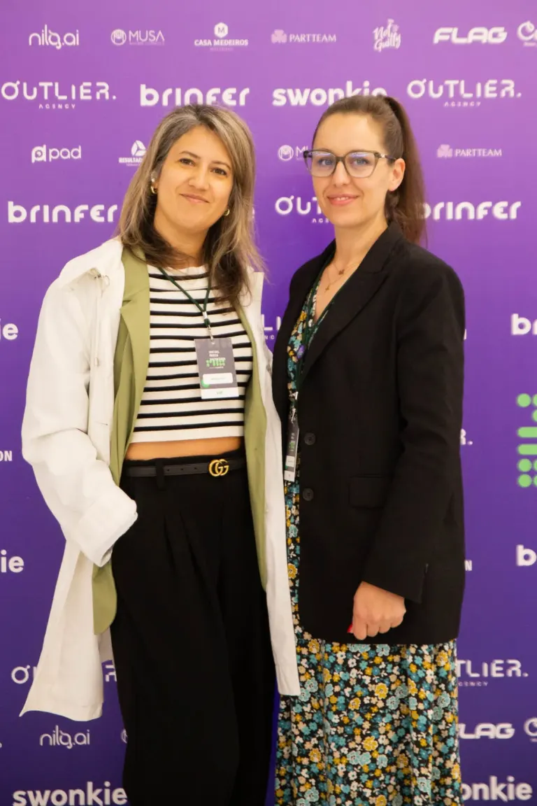 Duas mulheres posando para uma foto em um evento, com fundo roxo com logos de patrocinadores, uma usando jaqueta branca e...
