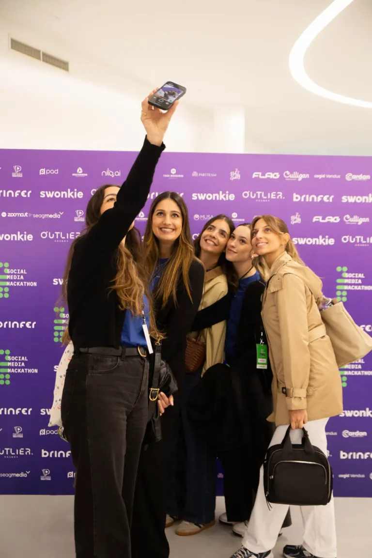 Grupo de cinco mulheres sorrindo, uma está tirando uma foto com o celular. Estão em um evento com fundo roxo com várias...
