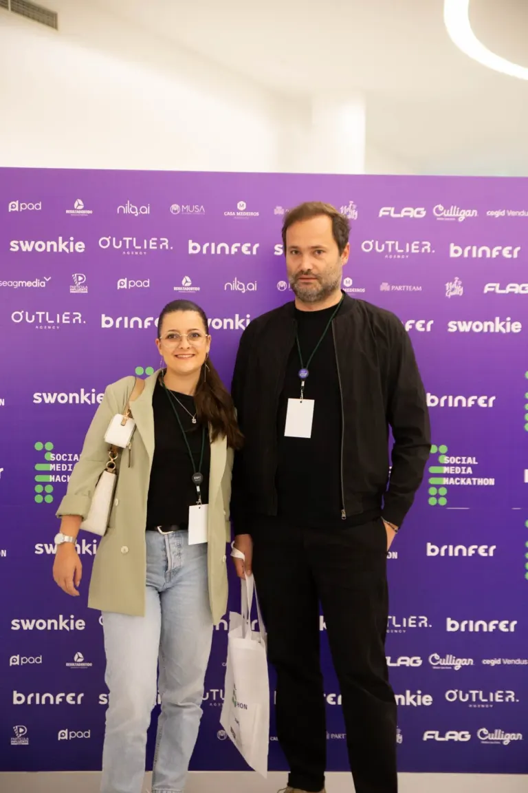 Homem e mulher posando para foto na conferência, com fundo roxo com logos de patrocinadores e marcas.