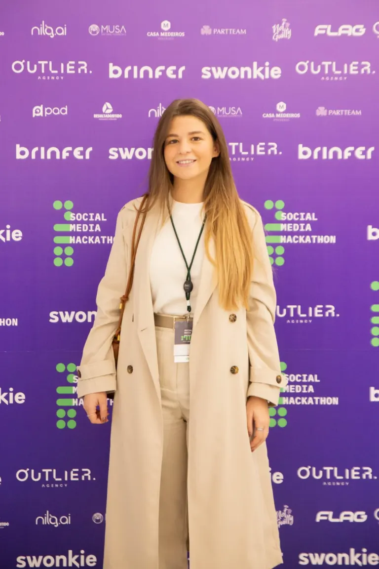 Mulher com cabelo castanho claro, vestindo roupa bege, de pé diante de fundo roxo com logotipos de patrocinadores e do...