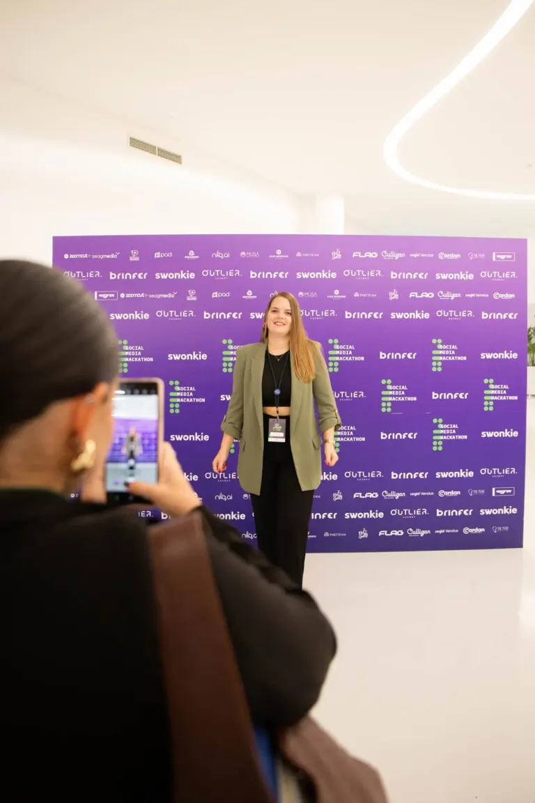 Pessoa posando para foto em evento, com banner roxo ao fundo contendo nomes e logotipos de marcas e patrocinadores.