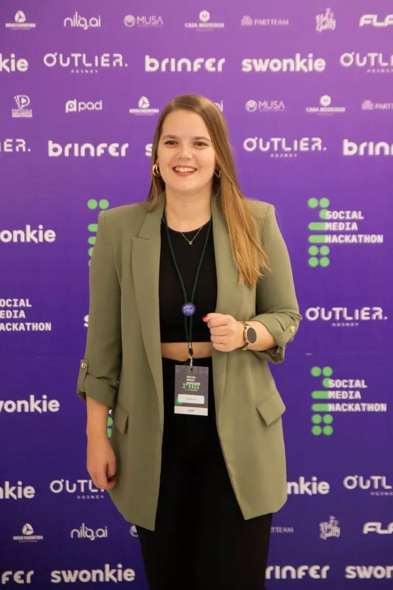 Mulher sorridente com cabelos longos, usando blazer verde claro, camiseta preta, brincos de argola dourados, pulseira, e...