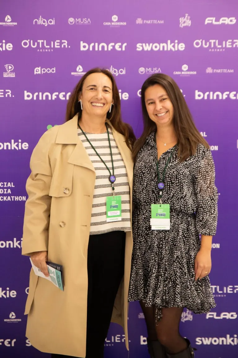 Duas mulheres posando para uma foto em um evento, ambas usando crachás verdes suspensos por tiras pretas. A mulher à...