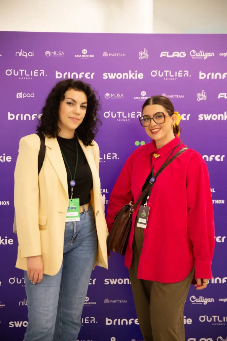 Duas mulheres com trajes casuais posando para uma foto em frente a um fundo roxo com logotipos de marcas e eventos.