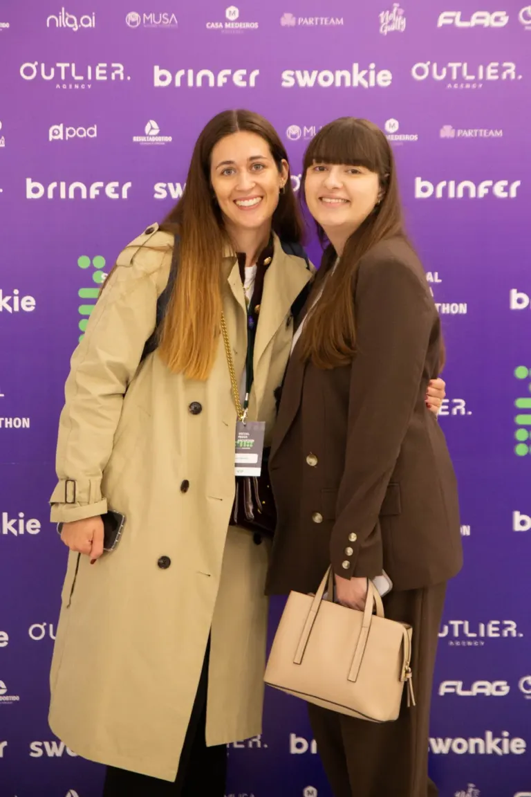 Duas mulheres sorridentes posando juntas para uma foto em evento, com fundo roxo e logotipos de patrocinadores