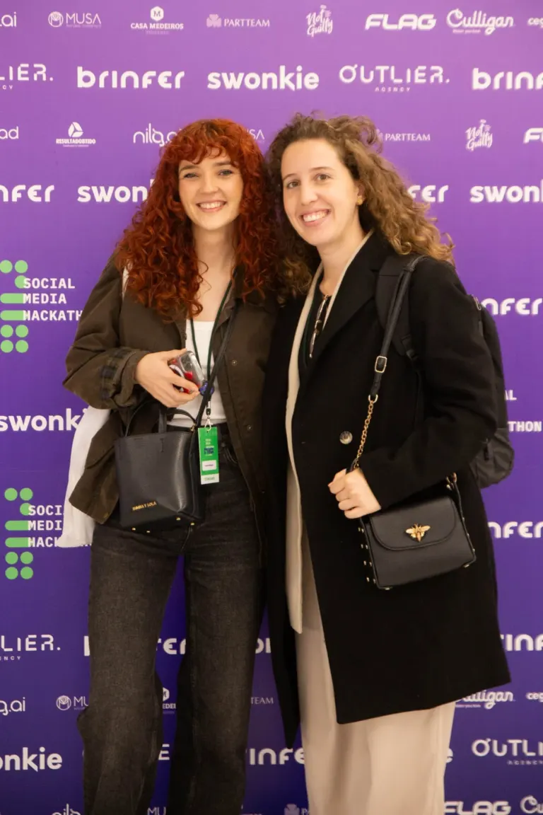 Duas mulheres sorrindo posando para foto em evento com fundo roxo com várias marcas e logos.