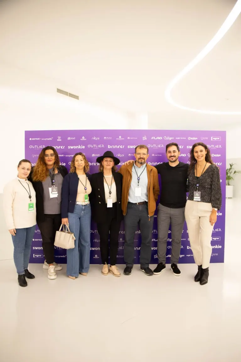 Grupo de oito pessoas posando para uma foto em uma conferência ou evento, com uma parede roxa ao fundo exibindo logotipos...