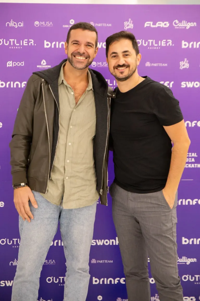 Dois homens sorrindo em evento com fundo roxo com logos de marcas e empresas.