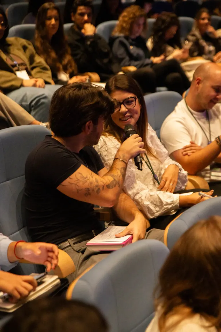 Pessoas sentadas em poltronas de audição, algumas conversando e outras usando celulares, em ambiente de teatro ou cinema.