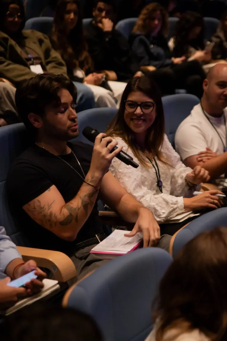 Pessoas sentadas em uma plateia, algumas usando microfones, observando atentamente a palestra ou evento em andamento, em...