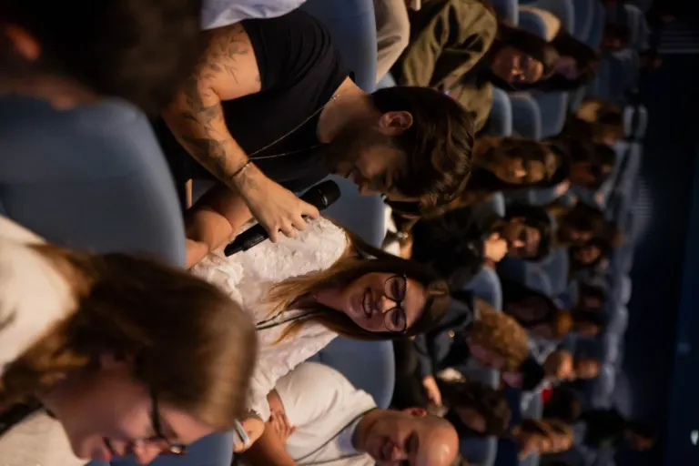 Pessoas sentadas em cadeiras de teatro ou cinema, assistindo a uma apresentação ou filme, com algumas sorrindo e outras...