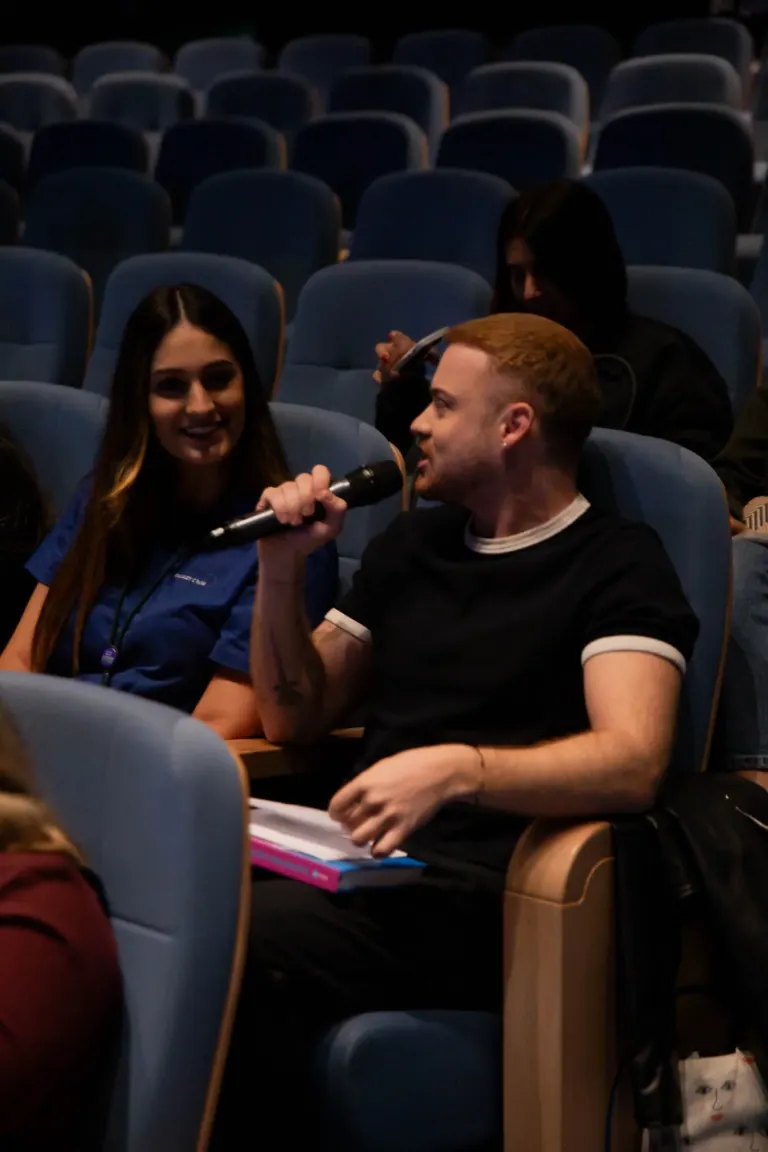 Pessoas conversando em uma sala de cinema ou teatro, uma delas segurando um microfone.