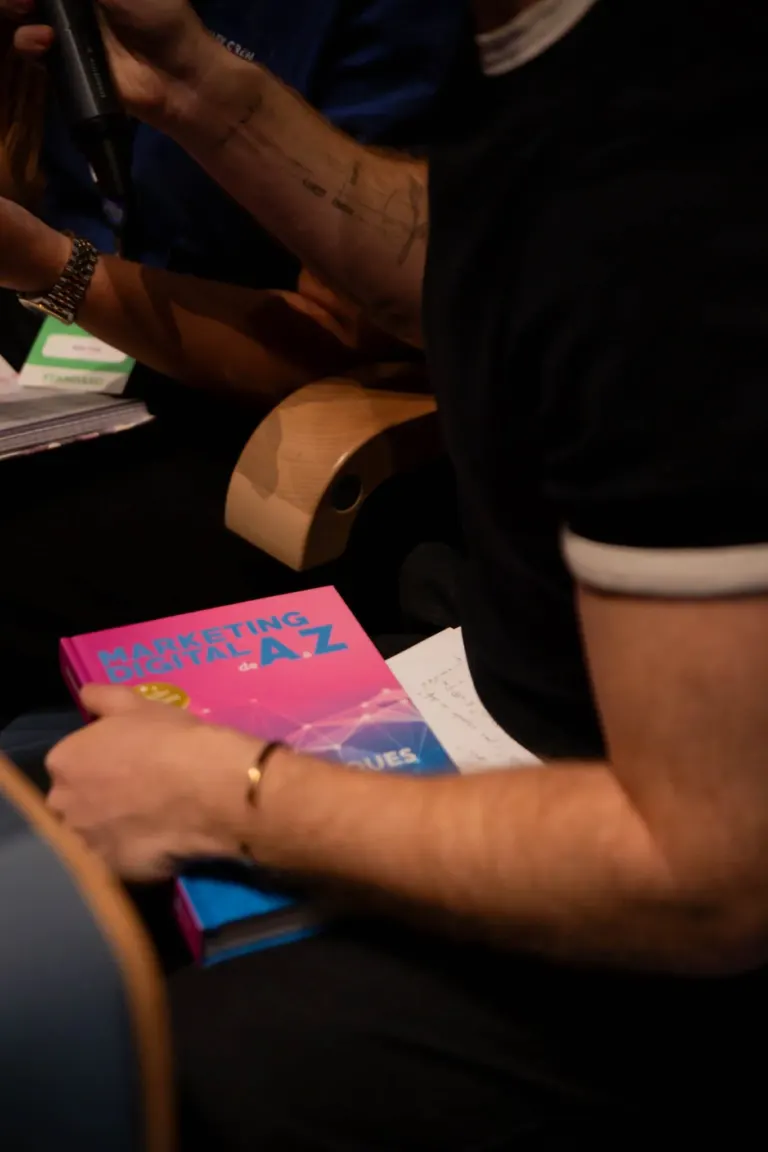 Pessoa segurando um livro com capa rosa intitulado 'MARKETING DIGITAL de A a Z' enquanto escreve em uma folha de papel,...