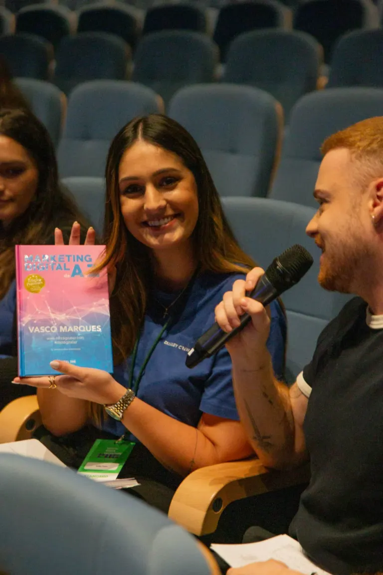 Mulher sorridente segurando um livro intitulado "Marketing Digital" ao lado de um homem que segura um microfone em um...