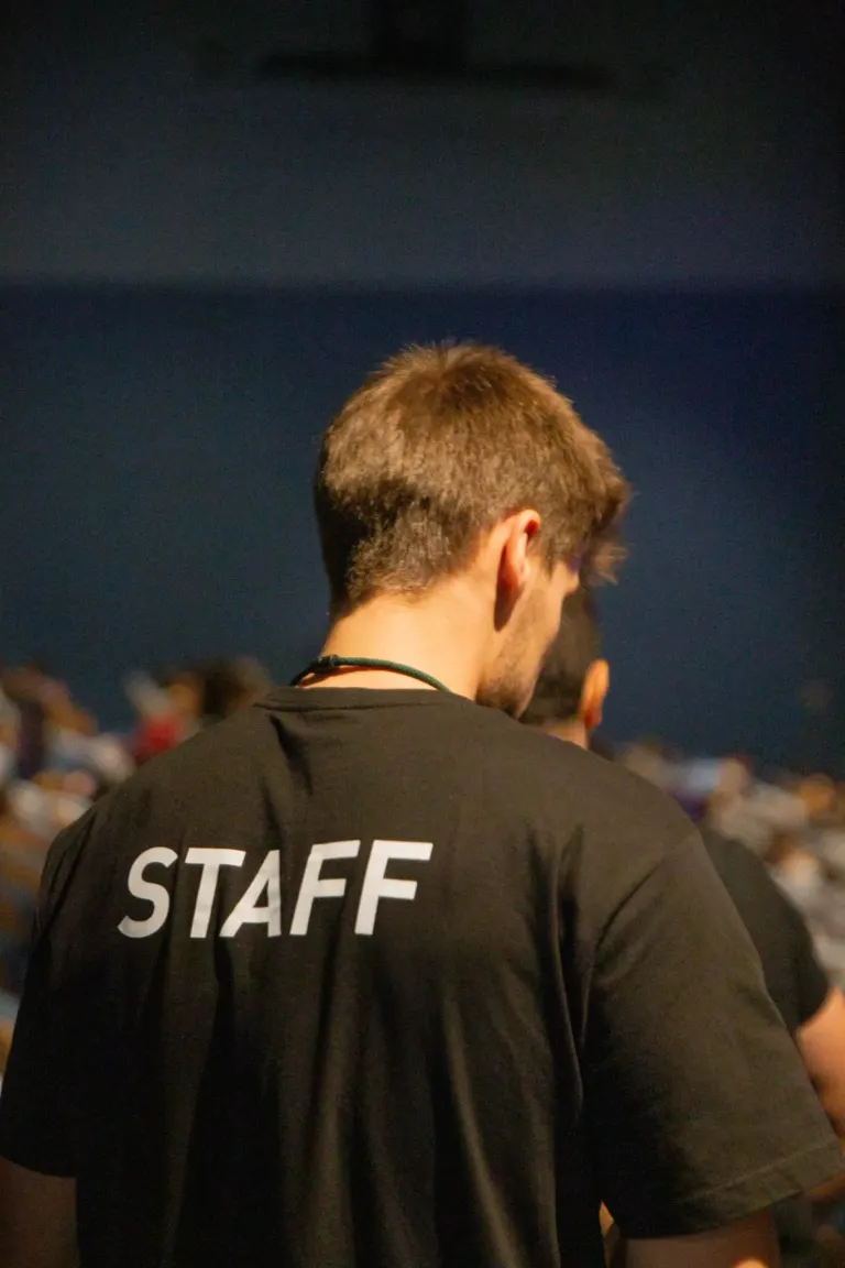 Homem com cabelo castanho claro e pele clara, vestindo uma camiseta preta com a palavra 'STAFF' na parte de trás, em um...