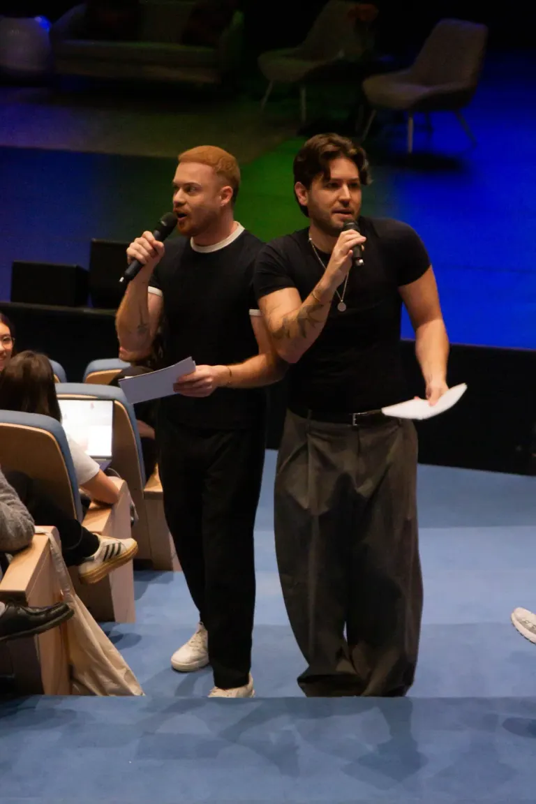 Dois homens em pé, segurando microfones, falando em um evento com assentos azuis e pessoas na plateia. Fundo escuro com...
