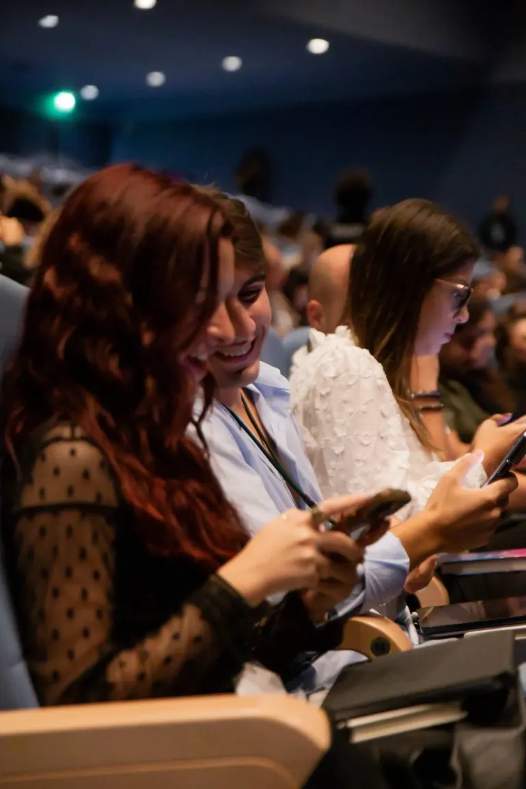Pessoas sentadas em poltronas de teatro ou auditório, com foco em três mulheres na frente que estão usando diferentes...