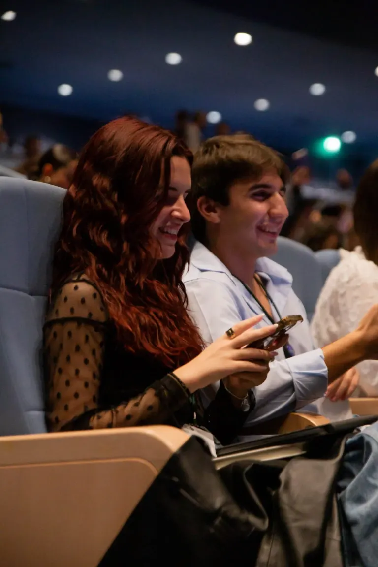 Homens e mulheres sentados em poltronas observando um evento, sorrindo e usando roupas casuais, em um ambiente de teatro...