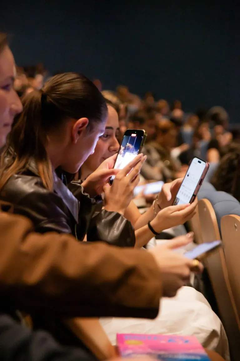 Pessoas sentadas em um teatro assistindo a uma apresentação, algumas usando celulares.