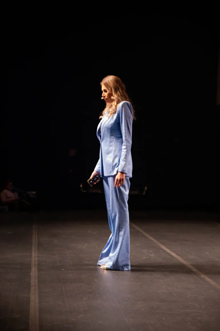 Mulher vestida com terno azul claro, com cabelo loiro, em pé no palco, segurando um controle remoto, com uma expressão...