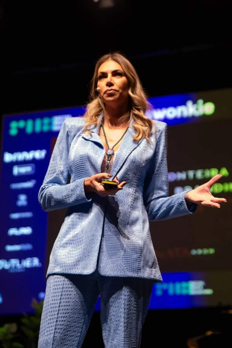 Mulher apresentando vestida com blazer azul xadrez, segurando um microfone e apresentando em palco com fundo digital.