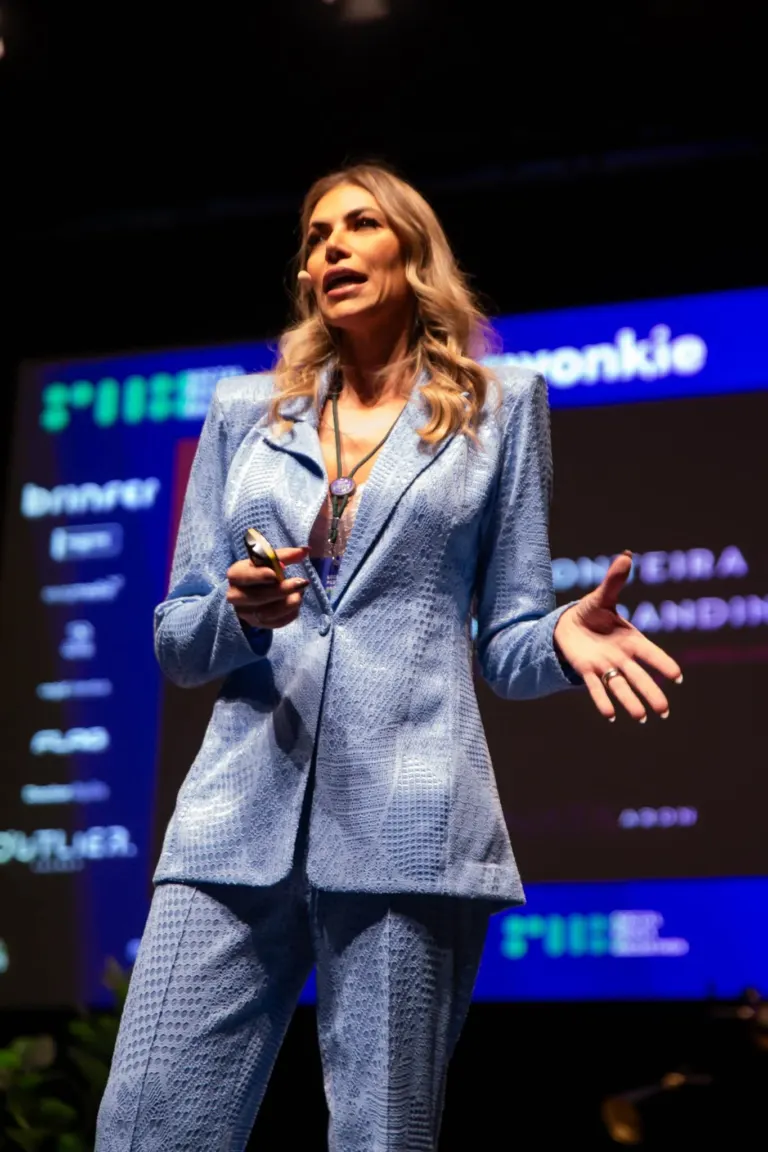 Mulher de cabelo louro claro, vestindo terno de tecido azul com padrão de texturas, falando em palco com um microfone de...