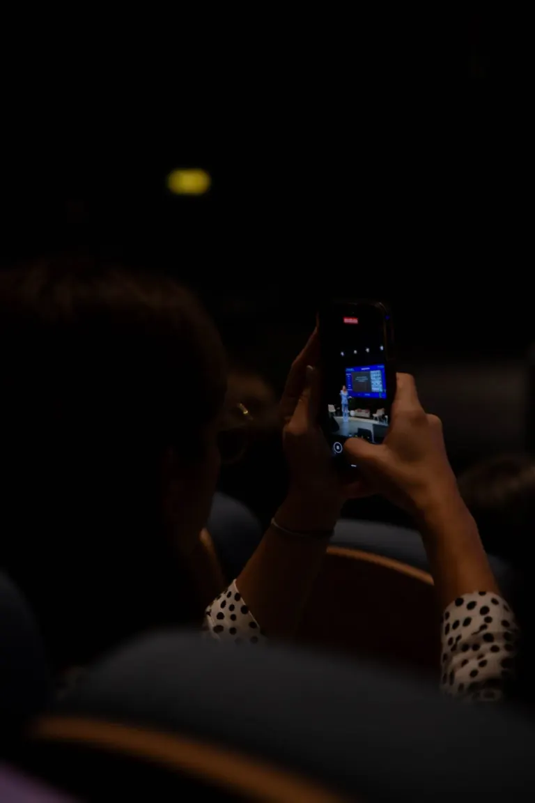 Pessoa segurando um smartphone para tirar uma foto de um palco com uma pessoa falando ao microfone, em um ambiente escuro.