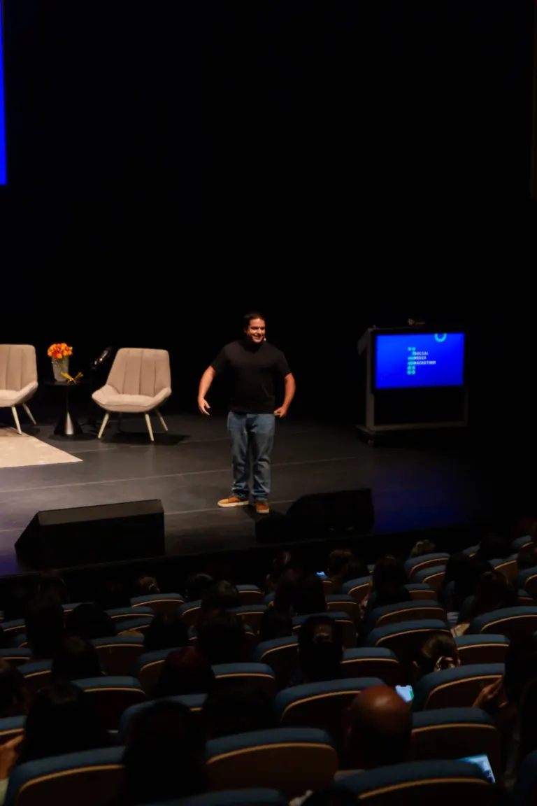 Cena de um palco de evento com um homem sorridente de camiseta preta e calça jeans, três cadeiras com uma mesa com vasos...