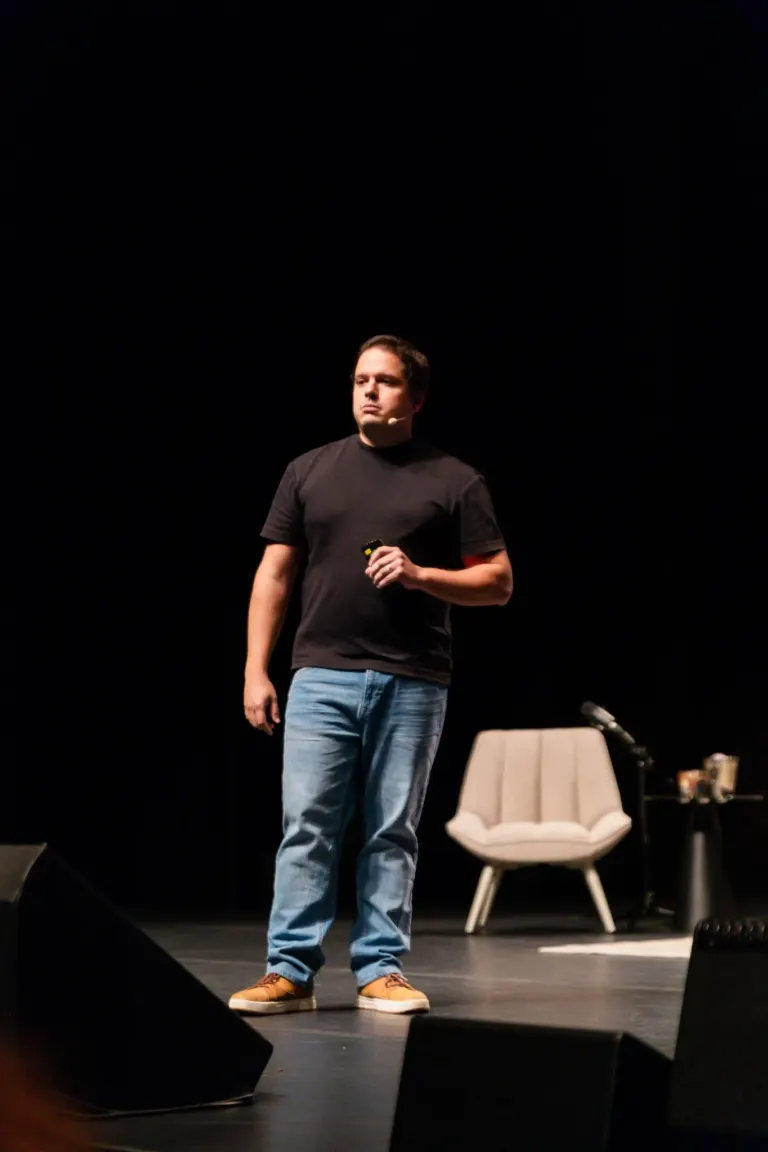 Homem de camiseta preta e jeans claro, de barba e cabelo escuro, segurando um controle remoto, em palco com cadeira...
