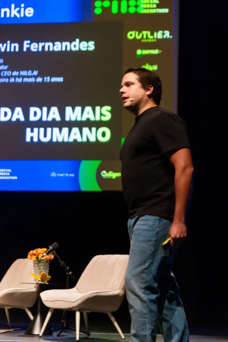 Homem de cabelo curto na fase de fala, de camiseta preta e calça jeans, de frente para uma tela de projeção com texto em...