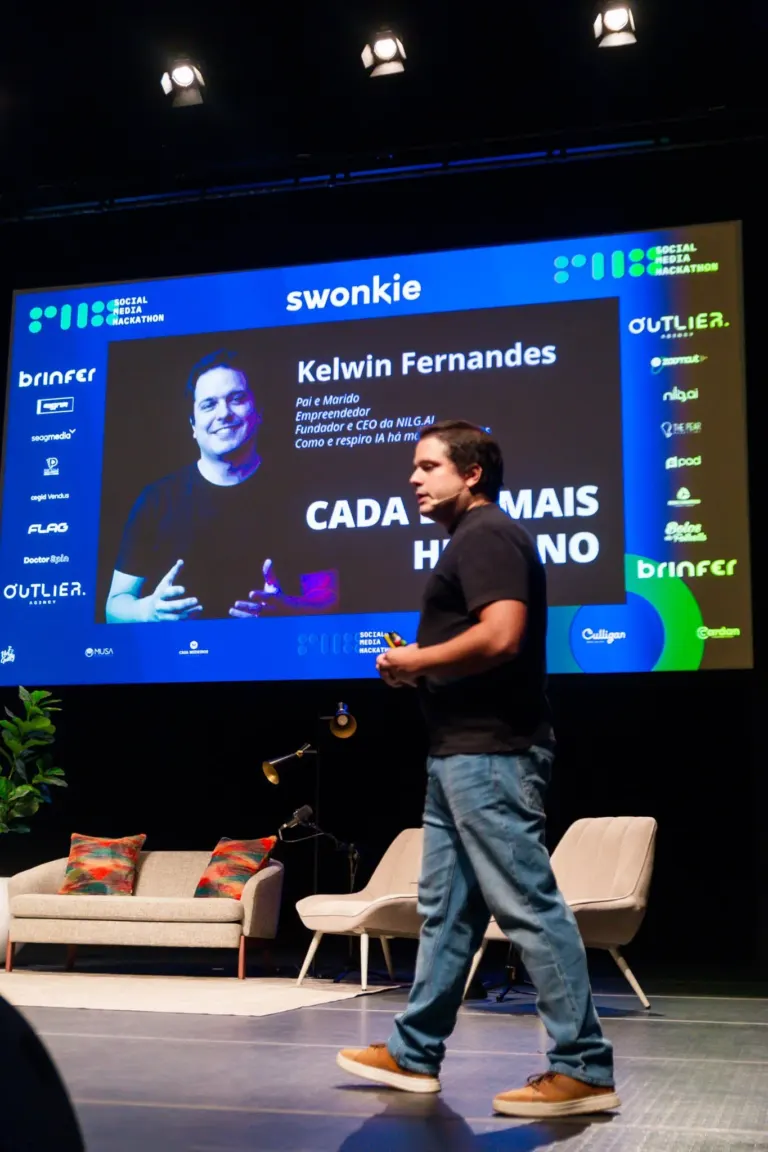 Homem de camiseta preta, calça jeans e sapatos brancos caminhando no palco de um evento chamado Social Media Hackathon,...