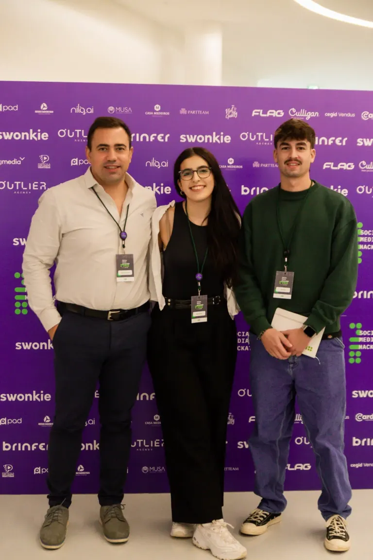 Três jovens em pé, sorrindo, na frente de um painel roxo com diversas logos e nomes de empresas, usando crachás de...