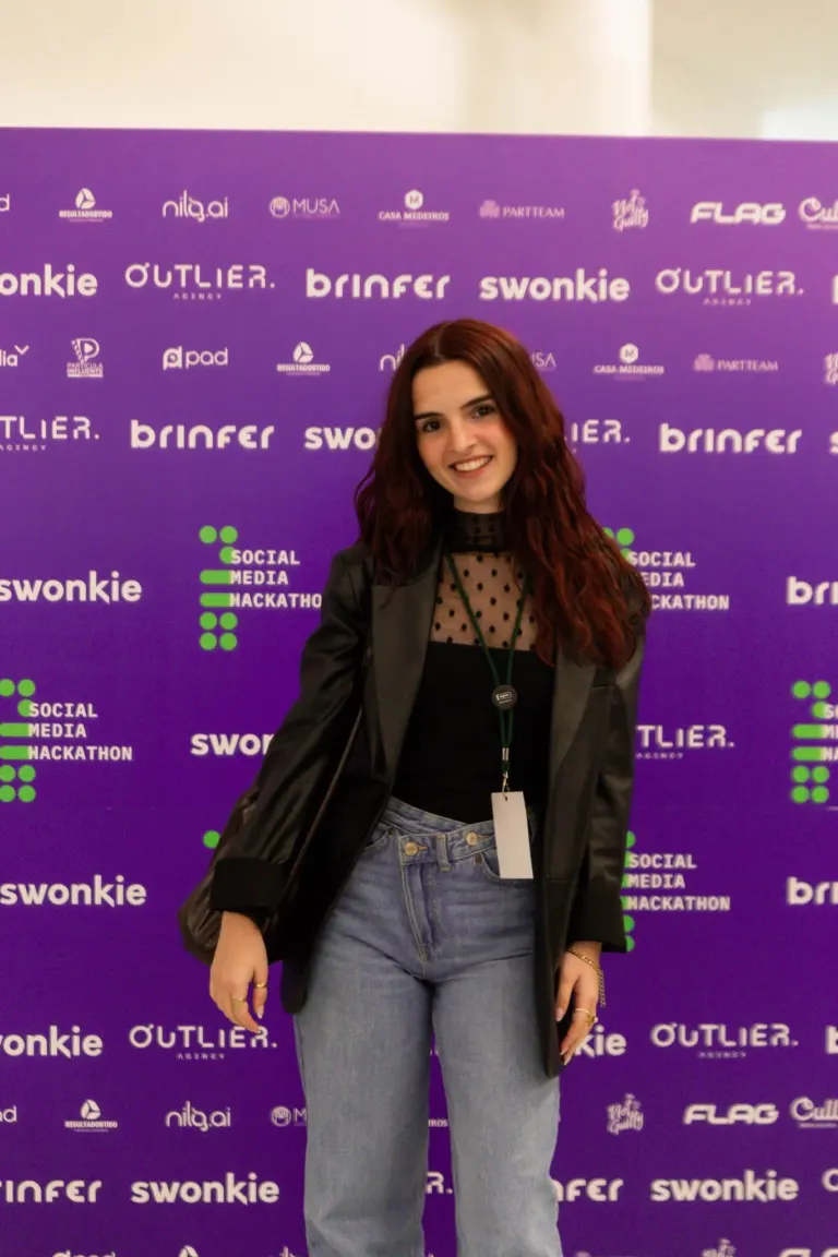 Mulher com cabelo castanho vestindo jaqueta preta, blusa preta com detalhes de tule e jeans, sorrindo na frente de um...