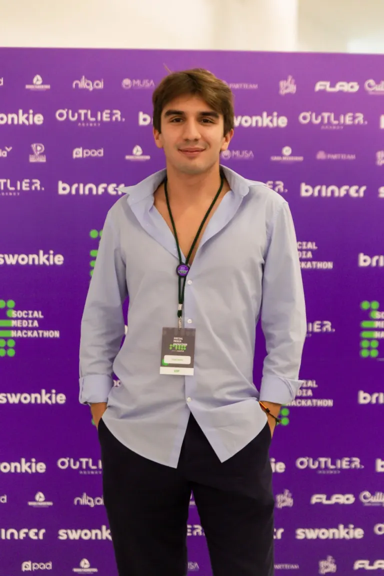 Jovem homem de cabelo castanho, vestindo camisa social cinza claro, de mangas arregaçadas, com um crachá de evento...