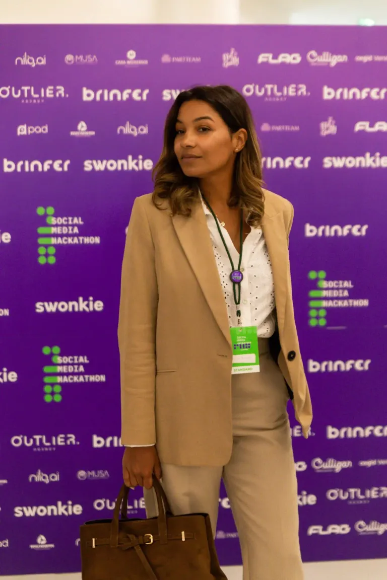 Mulher com cabelo castanho ondulado, vestindo blusa branca e blazer bege, segurando uma bolsa marrom, posando em frente a...