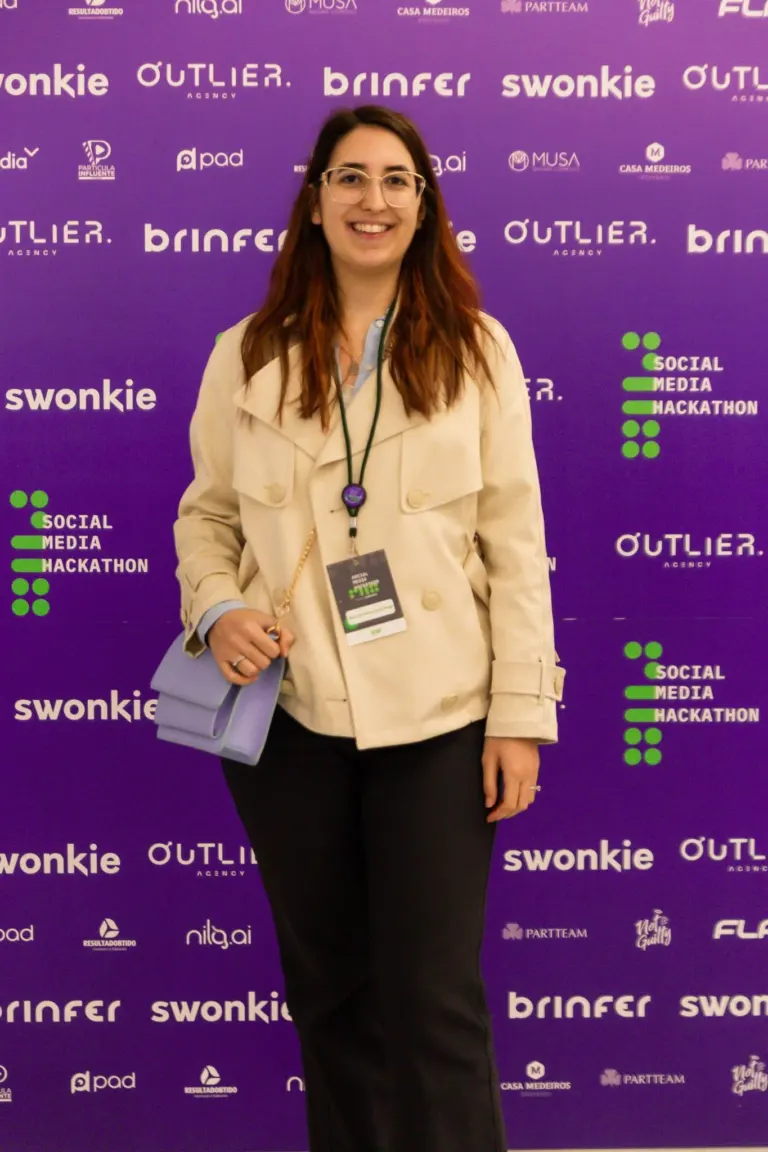 Mulher sorridente vestindo blusa azul e casaco bege, segurando uma bolsa lilás, em evento com fundo roxo com vários...