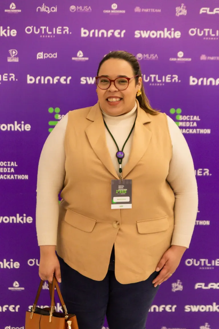Mulher sorridente de cabelos presos, usando óculos vermelhos, blusa branca de gola alta, colete bege, calça preta, com...